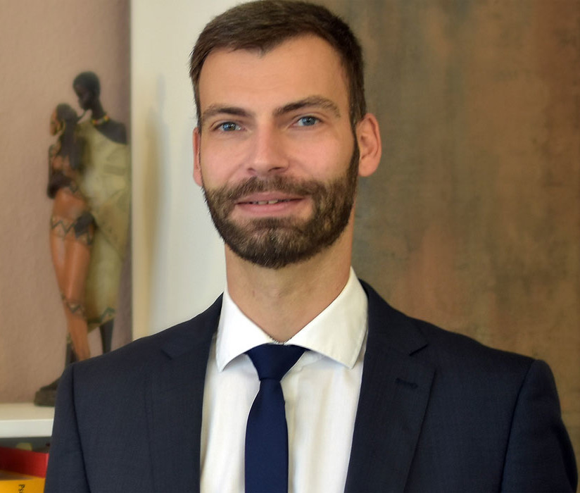 Ein Mann in Anzug vor einem Bücherregal mit Eulenfigur und Büchern.