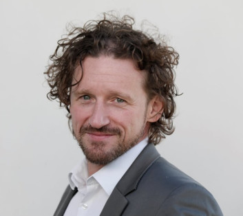 Porträt eines Mannes mit lockigem Haar und Bart, der einen schwarzen Anzug trägt.