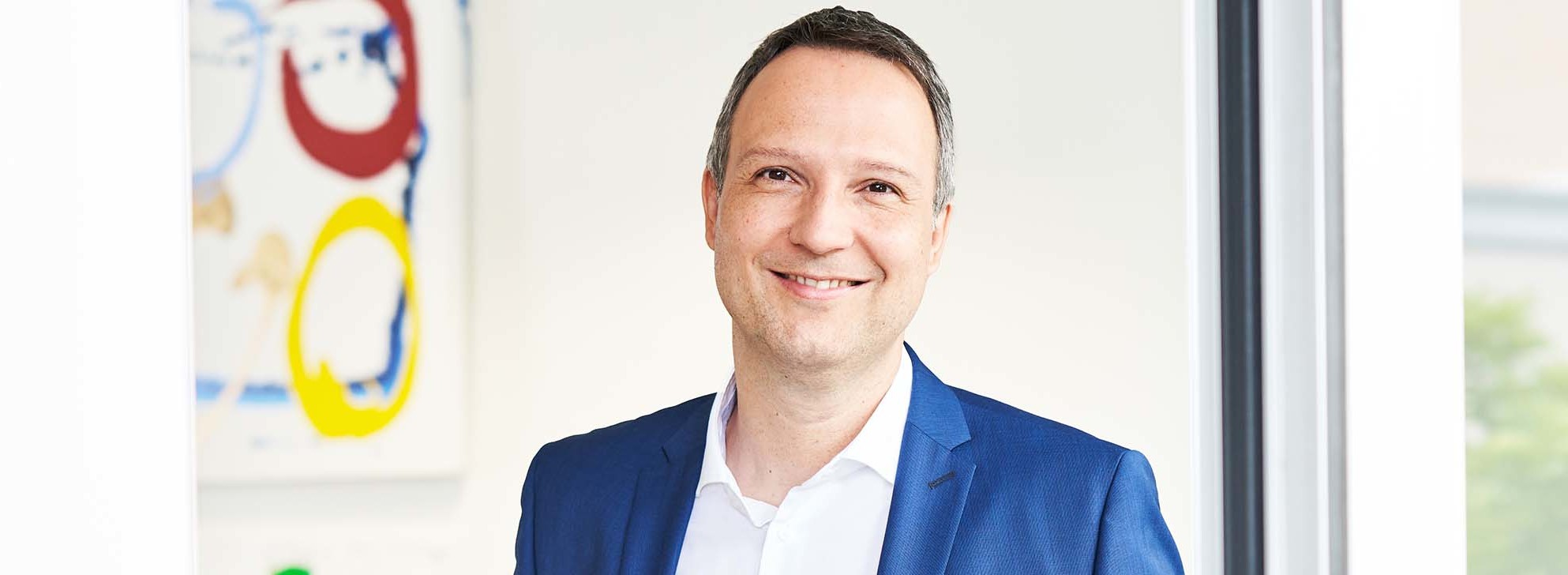 Mann in blauer Anzugjacke und weißem Hemd, lächelnd, stehend neben Fenster in modernem Büro.