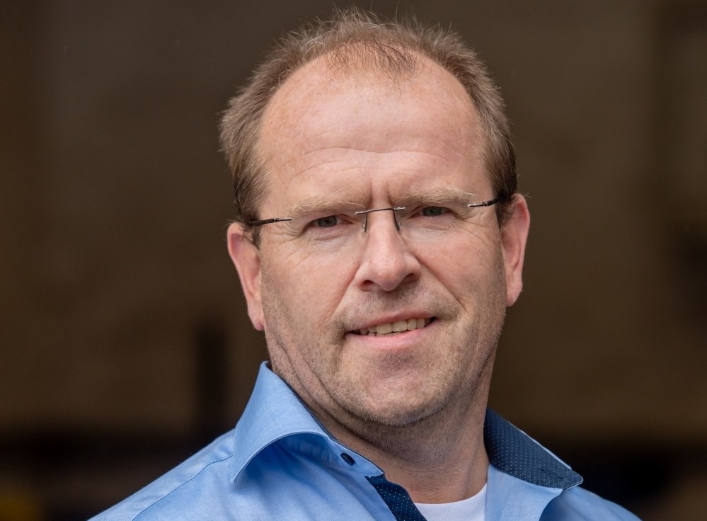 Mann mit Brille und blauer Hemd, lächelnd, im Freien.