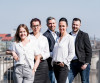 Eine Gruppe von fünf professionell gekleideten Menschen steht auf einer Brücke mit Stadtblick im Hintergrund.