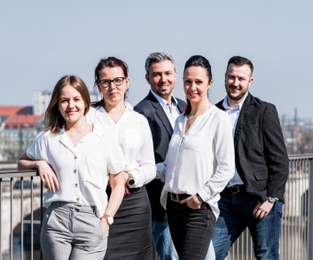 Eine Gruppe von fünf professionell gekleideten Menschen steht auf einer Brücke mit Stadtblick im Hintergrund.