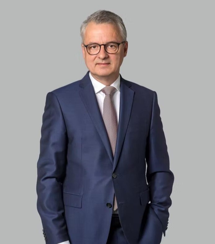 Ein Mann in einem dunklen Anzug mit Krawatte vor einem grauen Hintergrund.