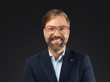 Porträt eines lächelnden Mannes in formeller Kleidung vor dunklem Hintergrund.