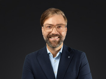 Porträt eines lächelnden Mannes in formeller Kleidung vor dunklem Hintergrund.