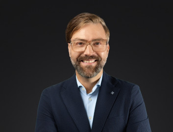 Porträt eines lächelnden Mannes in formeller Kleidung vor dunklem Hintergrund.