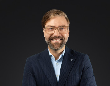 Porträt eines lächelnden Mannes in formeller Kleidung vor dunklem Hintergrund.