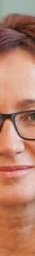 Porträt einer Frau mit roten Haaren, Brille, Perlenkette und schwarzem Blazer.
