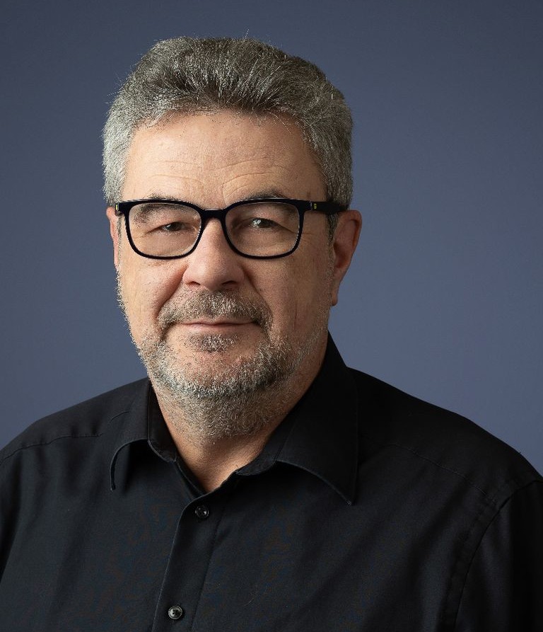 Porträt eines Mannes mit grauem Haar, Bart und Brille, trägt ein schwarzes Hemd vor einem dunklen Hintergrund.