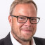 Porträt eines Mannes mit Brille, Anzug und weißem Hemd vor weißem Hintergrund.