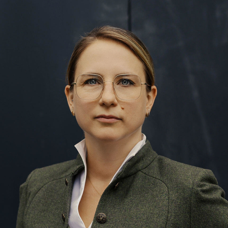 Frau mit Brille und grauer Jacke vor dunklem Hintergrund.