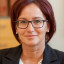 Porträt einer Frau mit roten Haaren, Brille, Perlenkette und schwarzem Blazer.