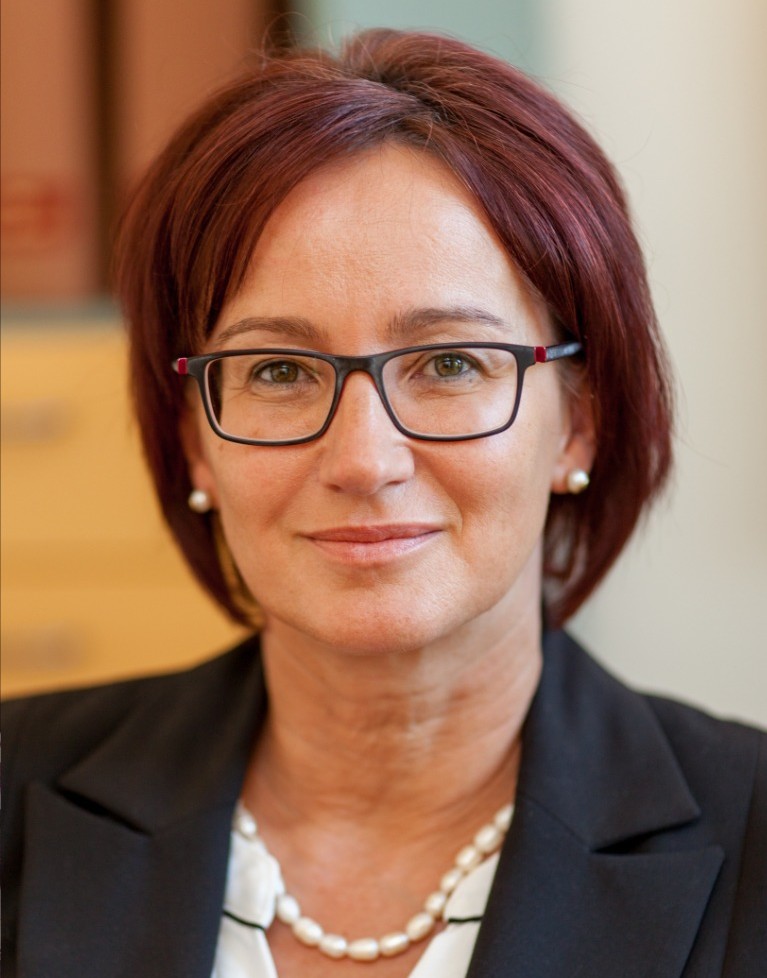 Porträt einer Frau mit roten Haaren, Brille, Perlenkette und schwarzem Blazer.