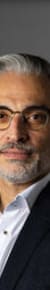Porträt eines Mannes mit grauem Haar und Brille, in formeller Kleidung, vor grauem Hintergrund.