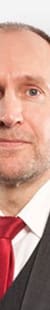 Ein Mann in einem dunklen Anzug mit roter Krawatte vor einem hellen Hintergrund.
