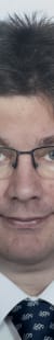 Porträt eines Mannes in formeller Kleidung mit Brille, dunklem Haar und einem freundlichen Lächeln.