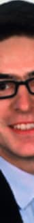 Porträt eines Mannes mit dunklem Haar, Brille und Anzug, lächelnd.