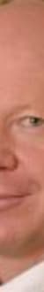 Ein Mann mit blassem Kopf und blauen Augen, trägt einen schwarzen Anzug und eine weiße Krawatte, im Hintergrund sind Bücherregale sichtbar.