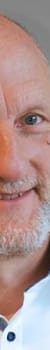 Ein lächelnder Mann mit kurzem, grauem Bart und blauen Augen, trägt ein weißes Hemd und steht vor einem grauen Hintergrund.