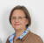Porträt einer Frau mit Brille, braunem Haar, blauer und weißer Bluse unter einem braunen Blazer vor weißem Hintergrund.