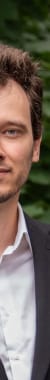 Junger Mann in formellem schwarzen Anzug vor grüner Hecke und Gebäudewand.