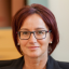 Porträt einer Frau mit roten Haaren, Brille und Perlenohrringen, lächelnd.