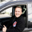 Lächelnder Mann im Auto, Daumen nach oben, trägt eine schwarze Jacke mit einem "Comes" Logo.