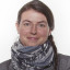 Porträt einer Frau mit braunen Haaren, die einen grauen Blazer und einen gemusterten Schal trägt.