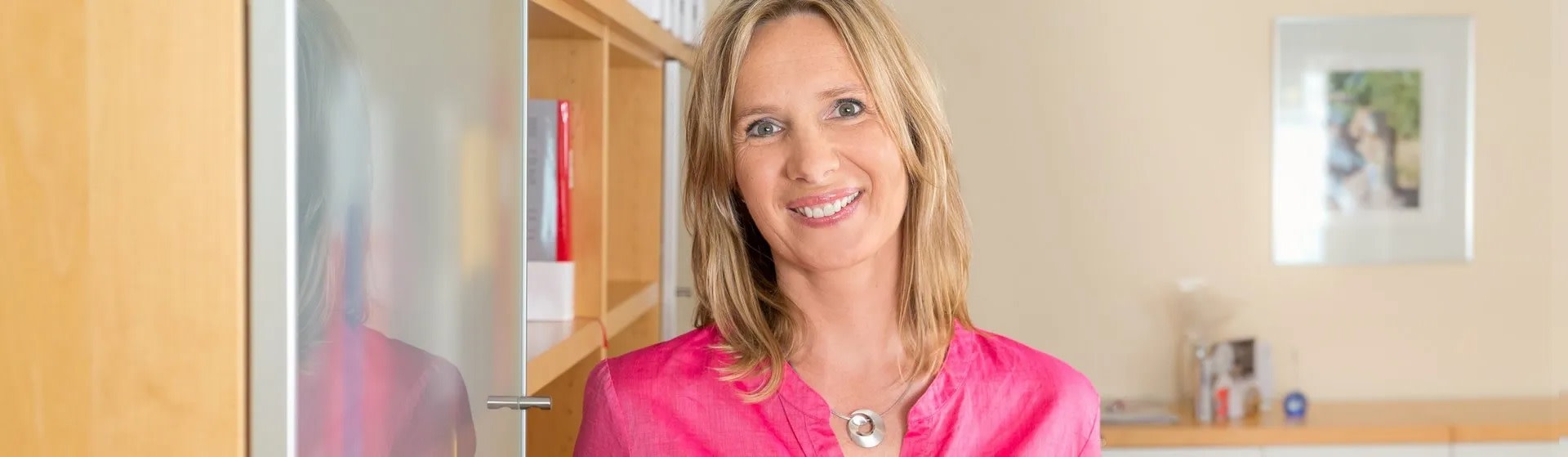 Lächelnde Frau in pinkem Hemd vor einem Bücherregal in einem Büro.