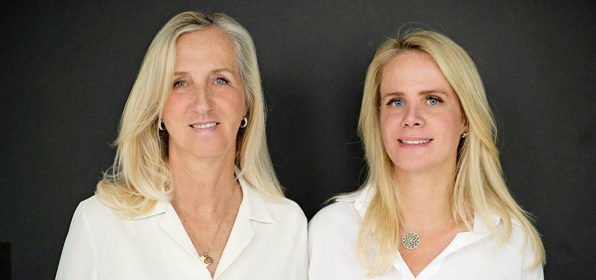 Zwei blonde Frauen in weißen Blusen vor dunklem Hintergrund.