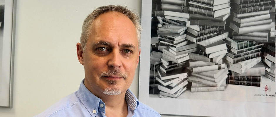 Mann mit grauem Haar und Bart vor einem Wandbild mit vielen Büchern.
