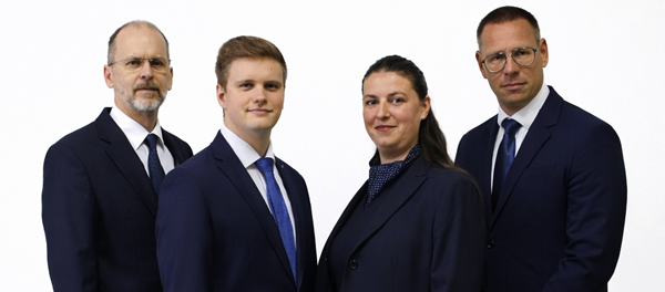 Vier professionell gekleidete Personen in dunklen Anzügen vor weißem Hintergrund.