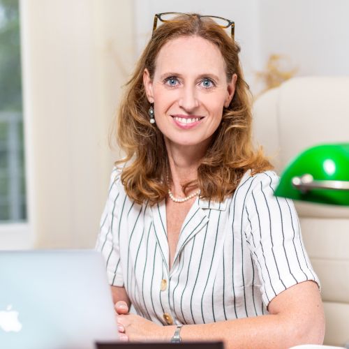 Frau sitzt an einem Schreibtisch mit einem Laptop, trägt eine weiße Bluse mit schwarzen Streifen und lächelt in die Kamera.