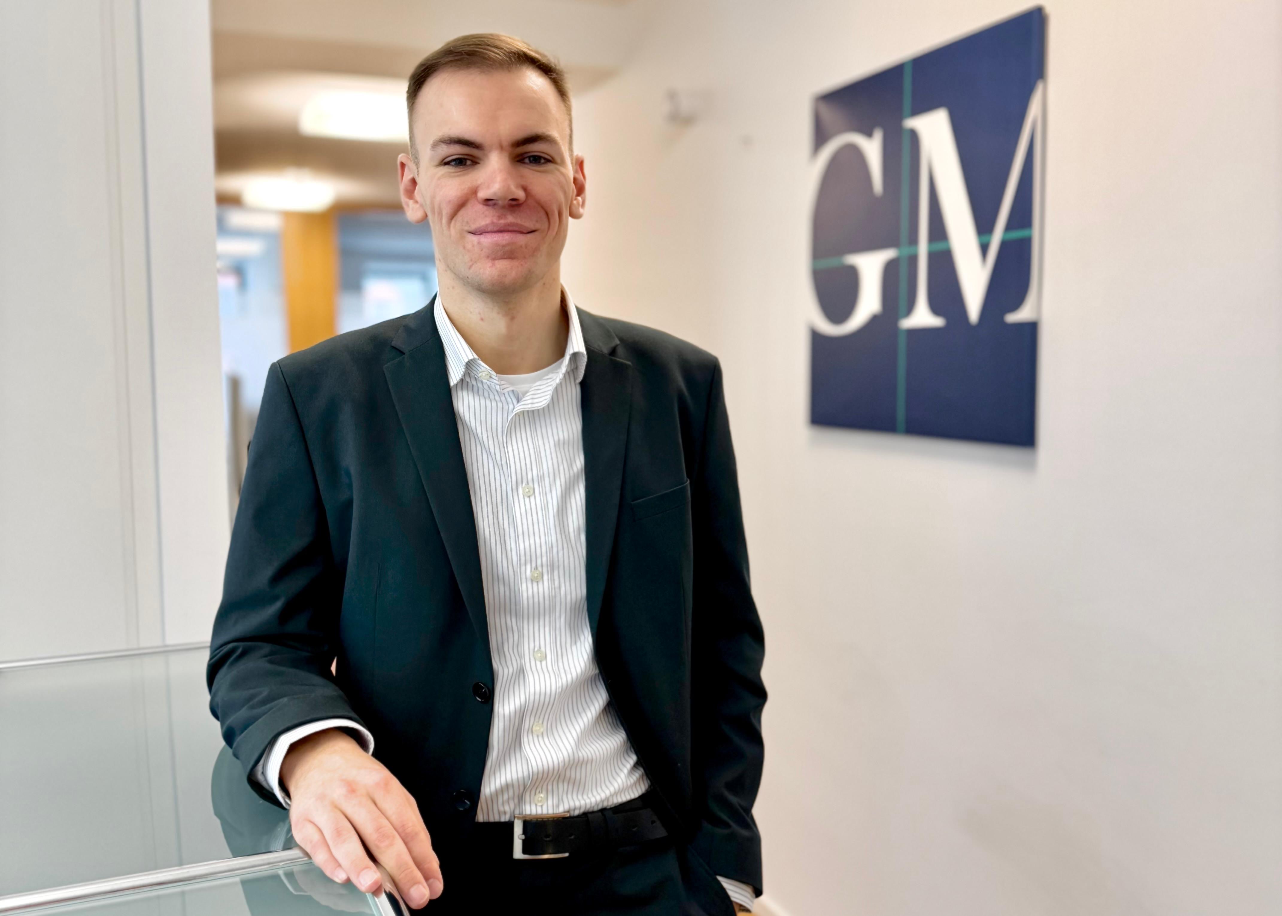 Junger Mann in Businesskleidung vor einem Firmenlogo an der Wand.