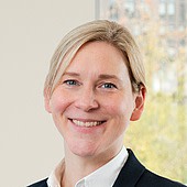 Das Bild zeigt eine lächelnde Frau mit blonden Haaren, die in einem hellen Raum vor einem Fenster steht.