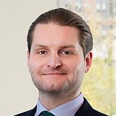 Ein Mann mit dunklem, glatt zurückgekämmtem Haar, trägt einen dunklen Anzug und ein weißes Hemd, lächelt leicht vor einem hellen Hintergrund.