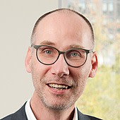 Ein Mann mit Brille, kurzhaarig, lächelnd, im Büro mit Fenster im Hintergrund.