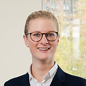 Ein Porträt einer jungen Frau mit Brille, die professionell gekleidet ist und vor einem Fenster steht.