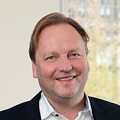 Ein Mann mit braunem Haar, freundlichem Lächeln, trägt einen dunklen Anzug und ein weißes Hemd.