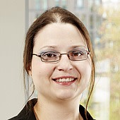 Porträt einer lächelnden Frau mit Brille vor einem unscharfen Hintergrund.