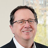 Ein Mann mit Brille, dunklem Haar und einem freundlichen Lächeln, trägt einen dunklen Anzug und eine weiße Hemd.