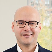 Ein Mann mit Brille, glattem Kopf und freundlichem Lächeln, trägt einen dunklen Anzug und eine weiße Hemd, im Hintergrund ein Fenster mit Blick auf eine Stadt.