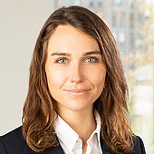 Ein Porträt einer jungen Frau mit braunen, gewellten Haaren, die einen schwarzen Blazer und ein weißes Hemd trägt.