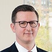 Ein Mann mit dunklem Haar, Brille und formeller Kleidung vor einem natürlichen Hintergrund.