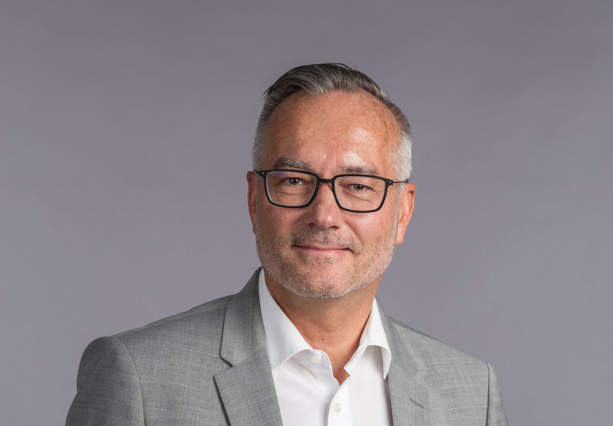 Porträt eines Mannes in einem grauen Anzug und weißen Hemd, mit Brille, vor einem grauen Hintergrund.