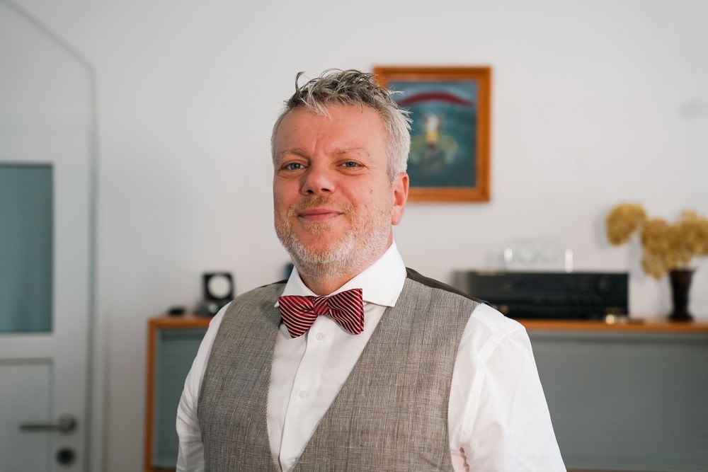 Mann mit grauem Bart und grauem Haar, trägt weiße Hemd, grauen Weste und roten Fliege, steht in einem Raum mit moderner Einrichtung.