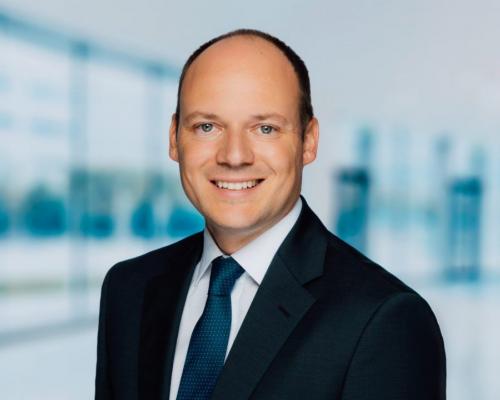 Ein Mann in einem dunklen Anzug und Krawatte lächelt vor einem modernen Bürogebäude.
