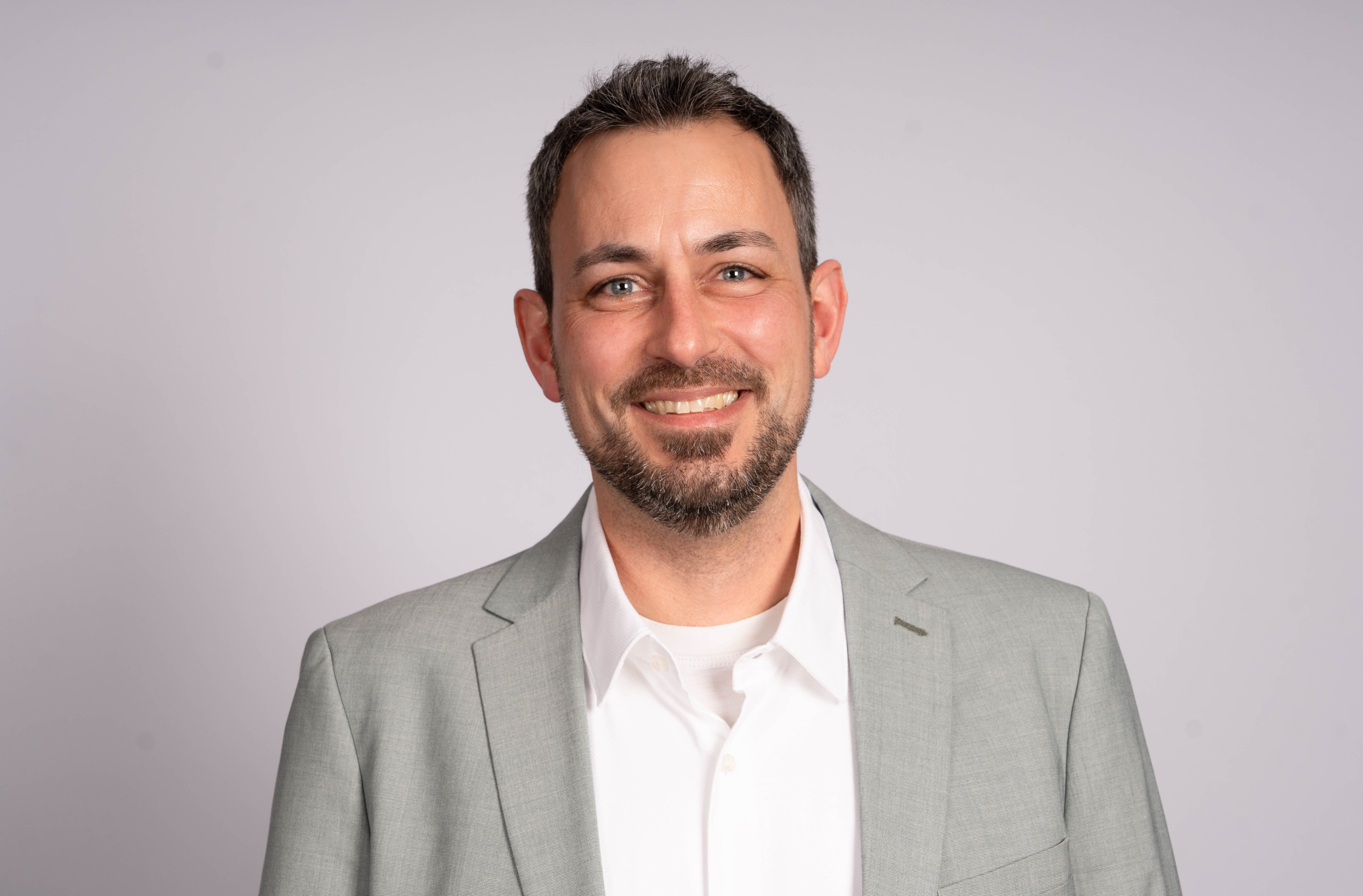 Lächelnder Mann in einem hellgrauen Anzug und weißem Hemd vor neutralem Hintergrund.