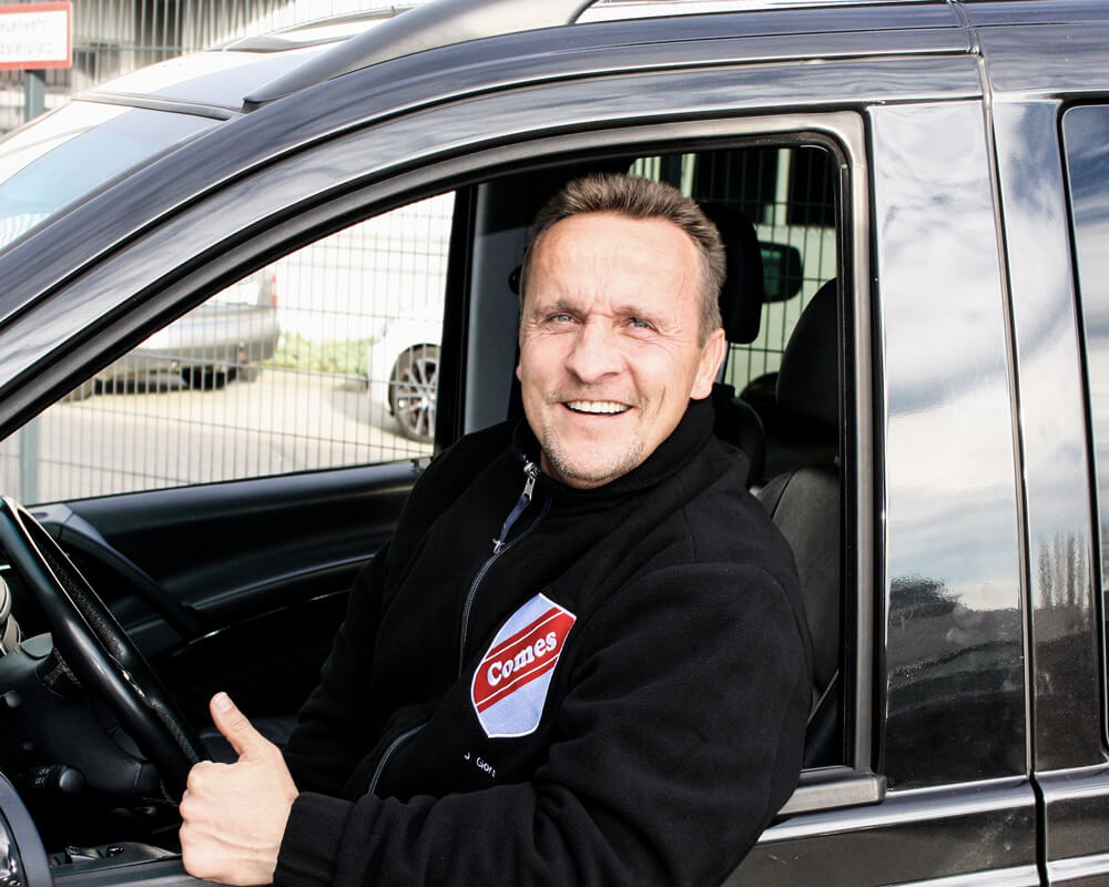 Lächelnder Mann im Auto, Daumen nach oben, trägt eine schwarze Jacke mit einem "Comes" Logo.