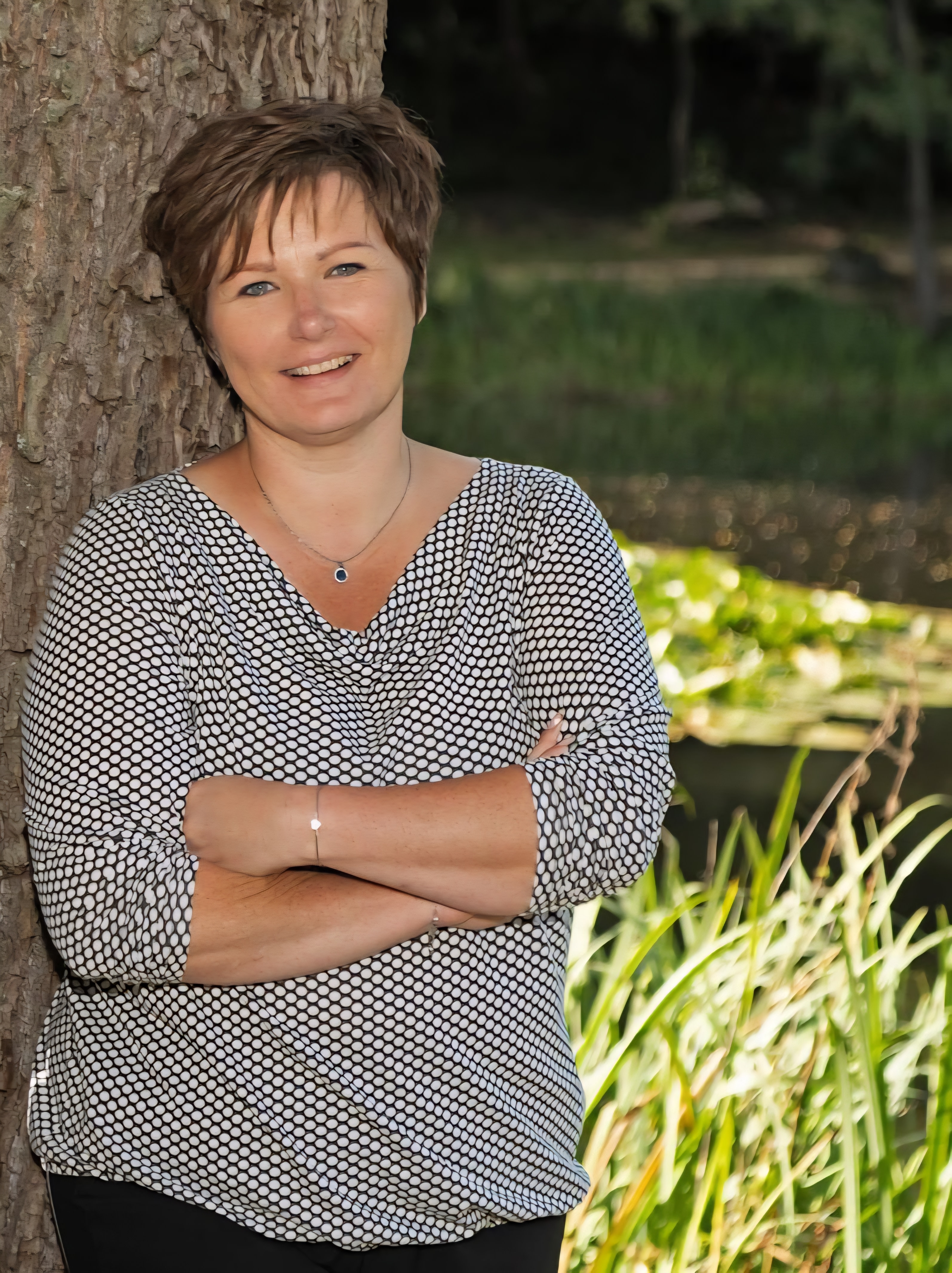 Frau lehnt an Baum am Wasser im Freien, lächelnd.
